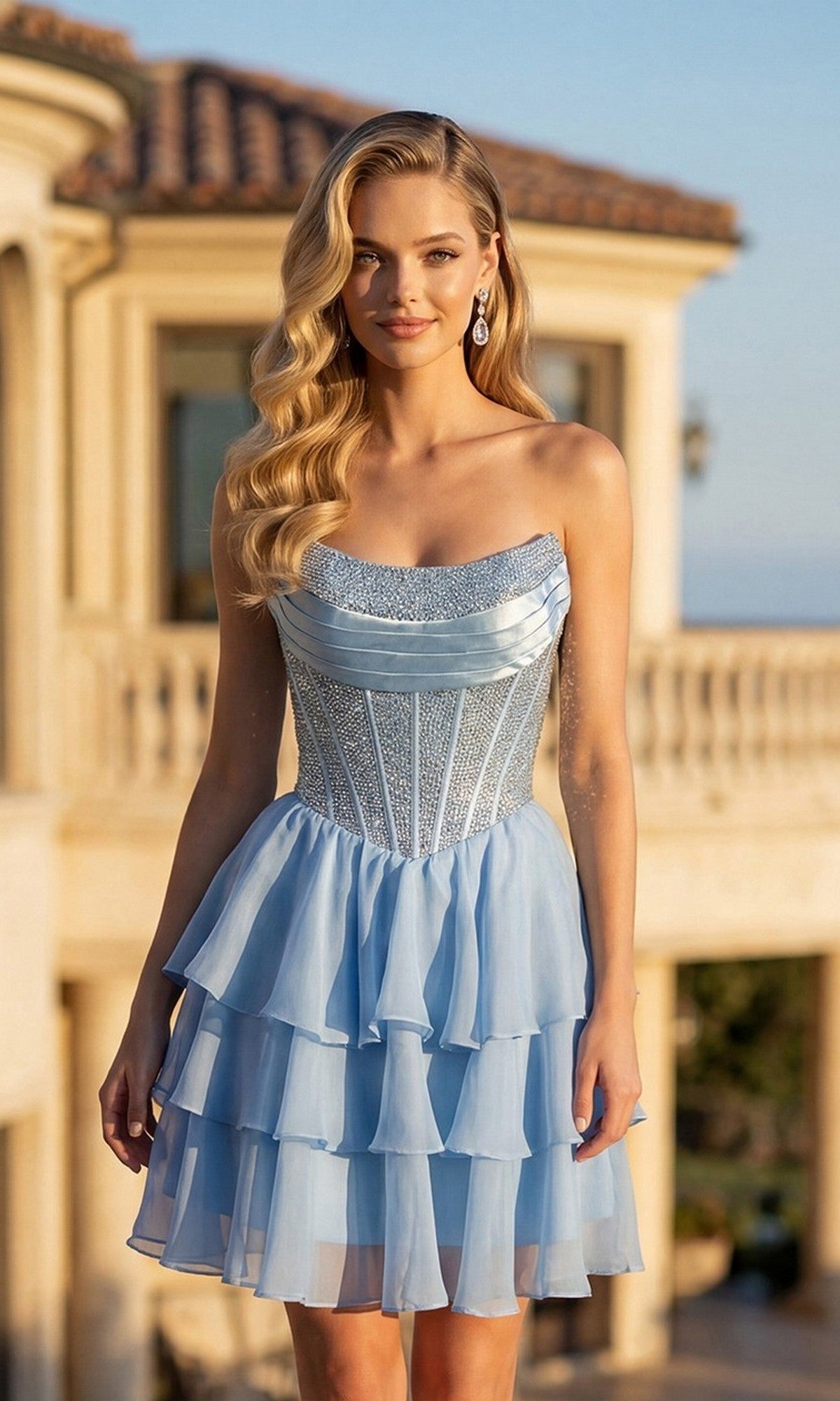 A woman stands outdoors in front of a columned building, wearing the Juliet JT7135S short homecoming dress in sky blue, featuring a strapless sequined corset bodice and a light blue chiffon skirt.